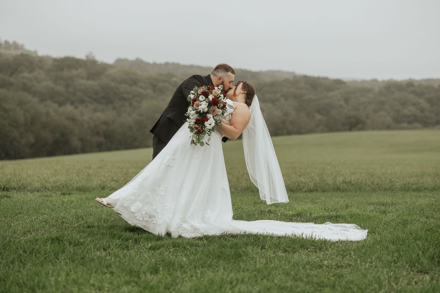 A couple shares a romantic embrace in white wedding attire against a scenic green hillside landscape.