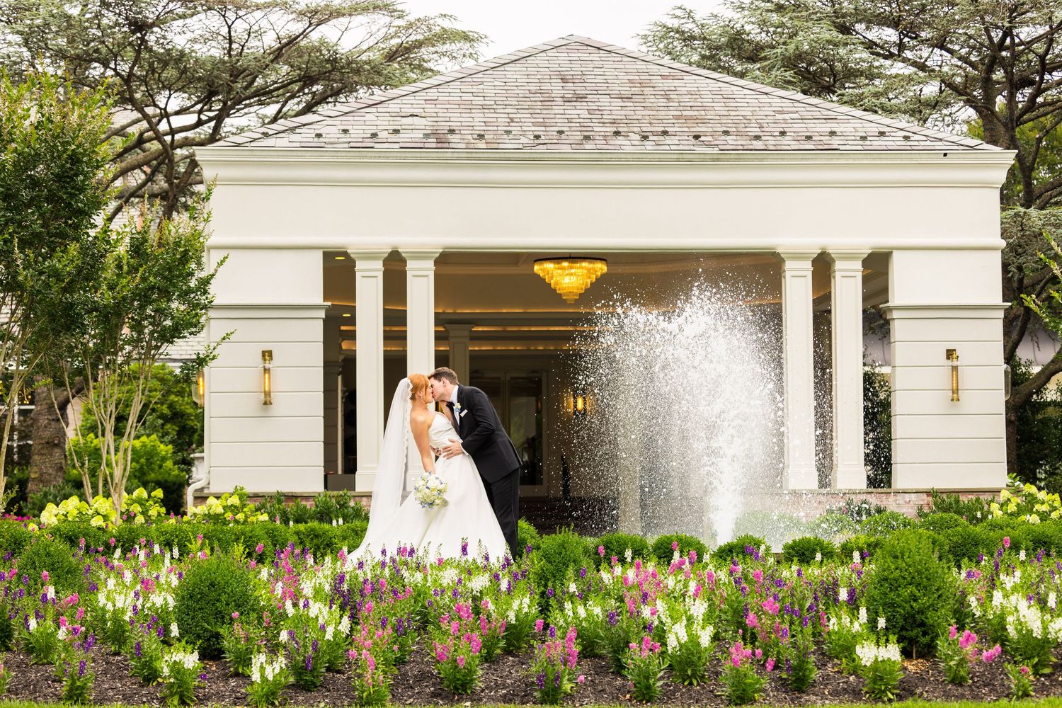 Elegant white gazebo with pillared entrance and flowing fountain surrounded by colorful garden flowers.