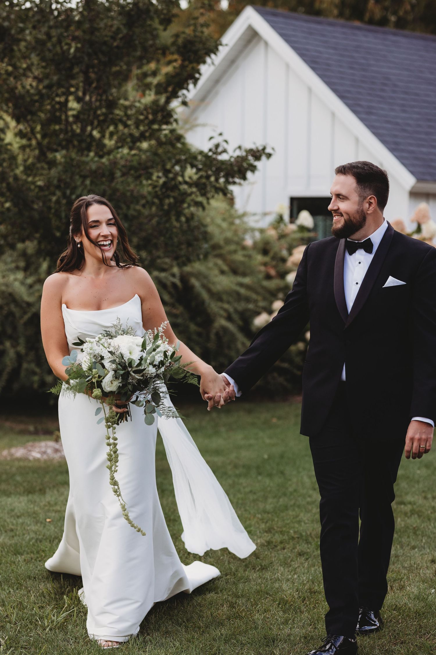 A wedding couple walks together on a lawn outside a white church, wearing elegant white dress and black tuxedo.