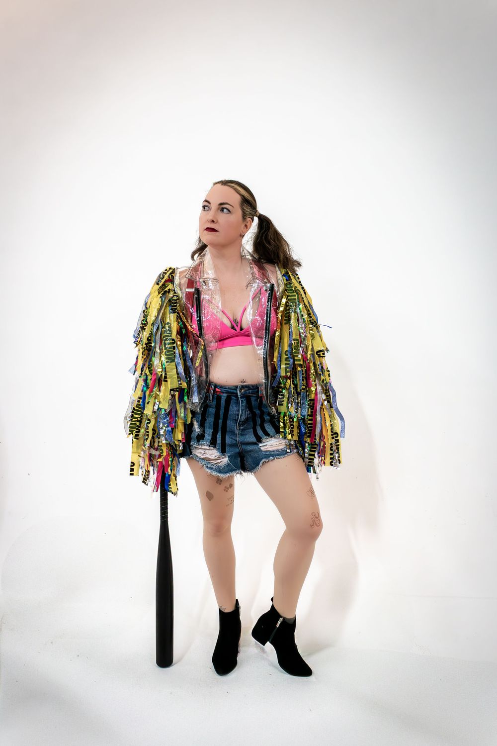Performer in denim shorts and colorful fringe jacket poses dynamically against white background.