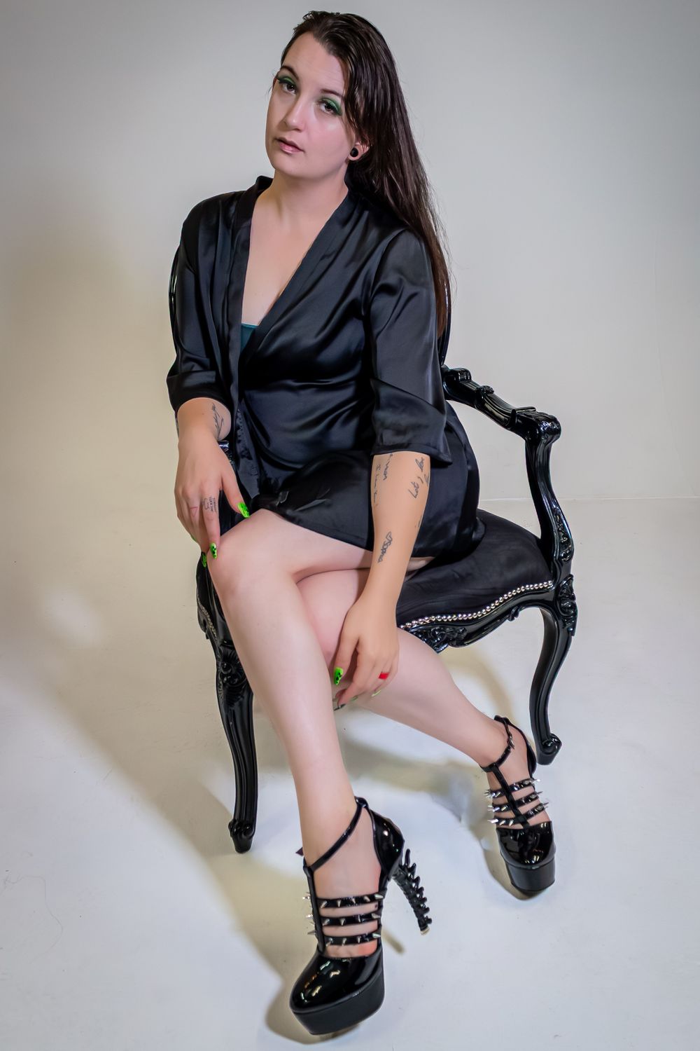 Person wearing black satin top and heels poses on dark chair in studio setting.