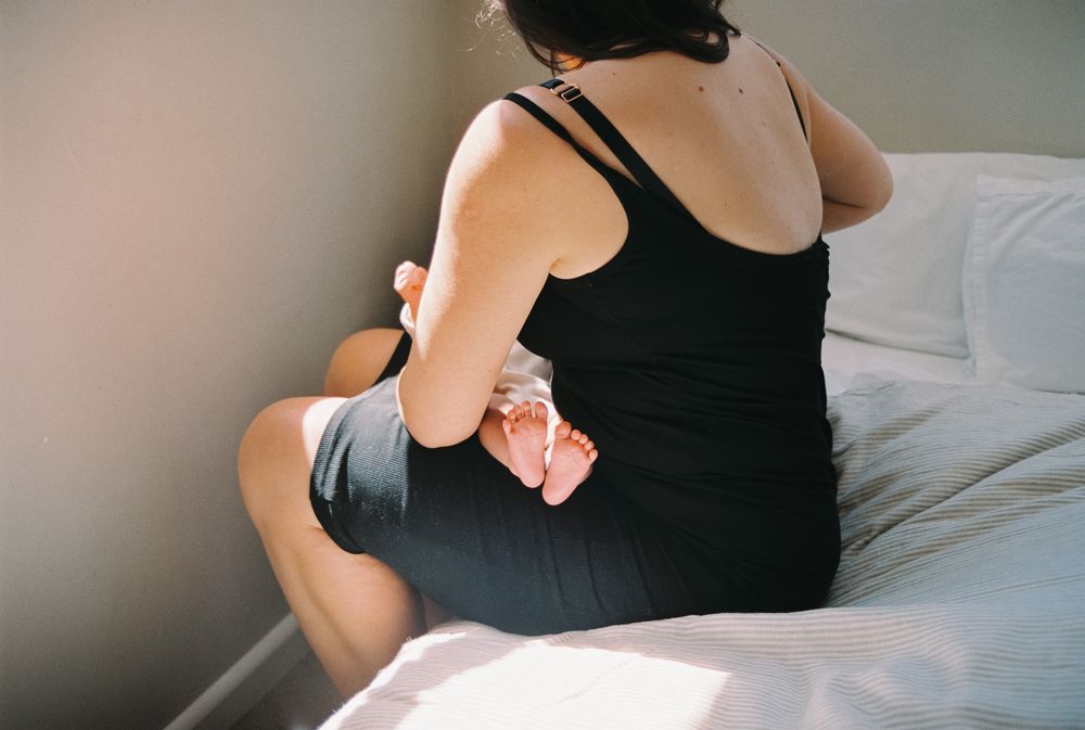 A woman in a black dress stands by a window looking down at a newborn baby in her arms.