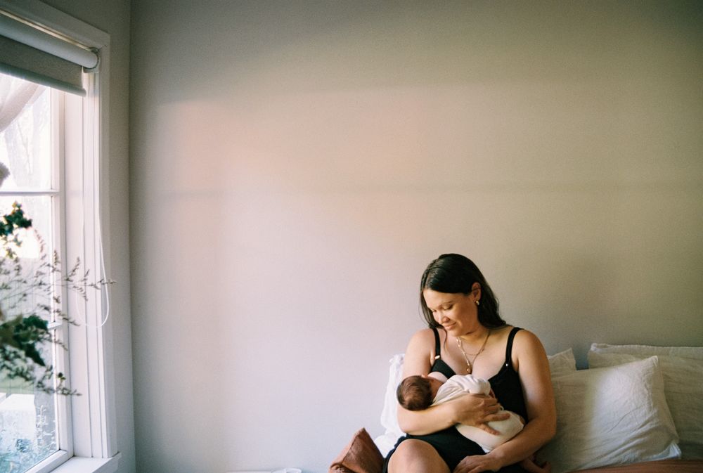 A tender moment of mother nursing baby by a sunny window in a peaceful bedroom setting.