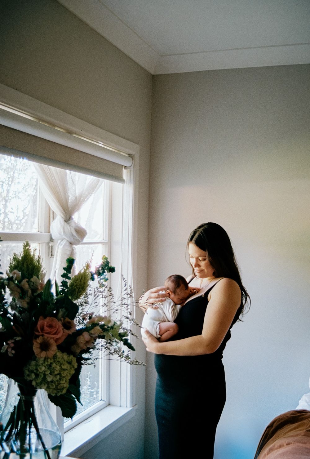 Five tender moments captured of a woman in black holding a newborn baby near a bright window with flowers.