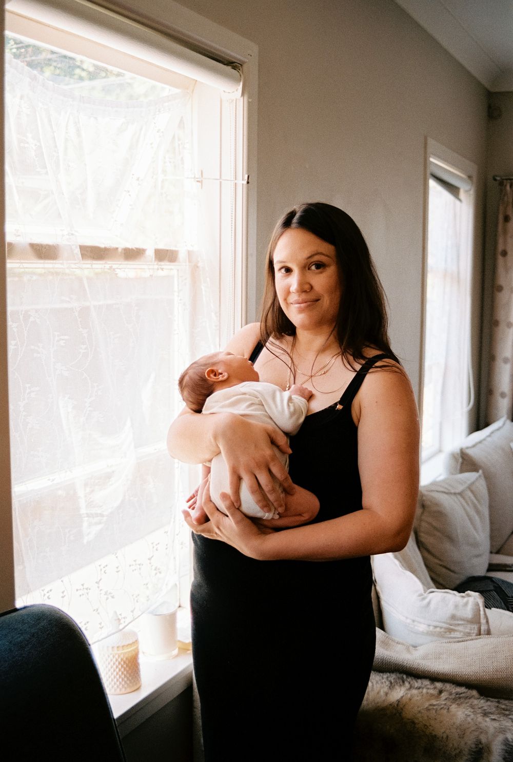 A tender moment between parent and newborn near sunlit windows in a cozy bedroom setting.