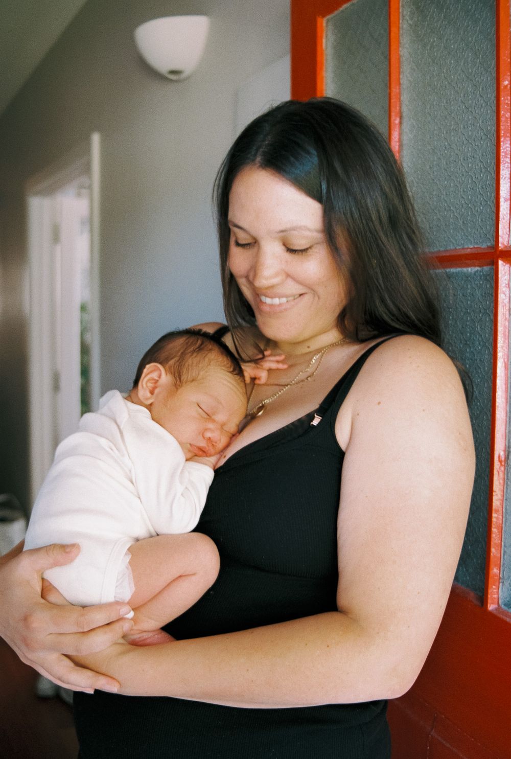 A tender moment between mother and sleeping baby near a window with orange trim.