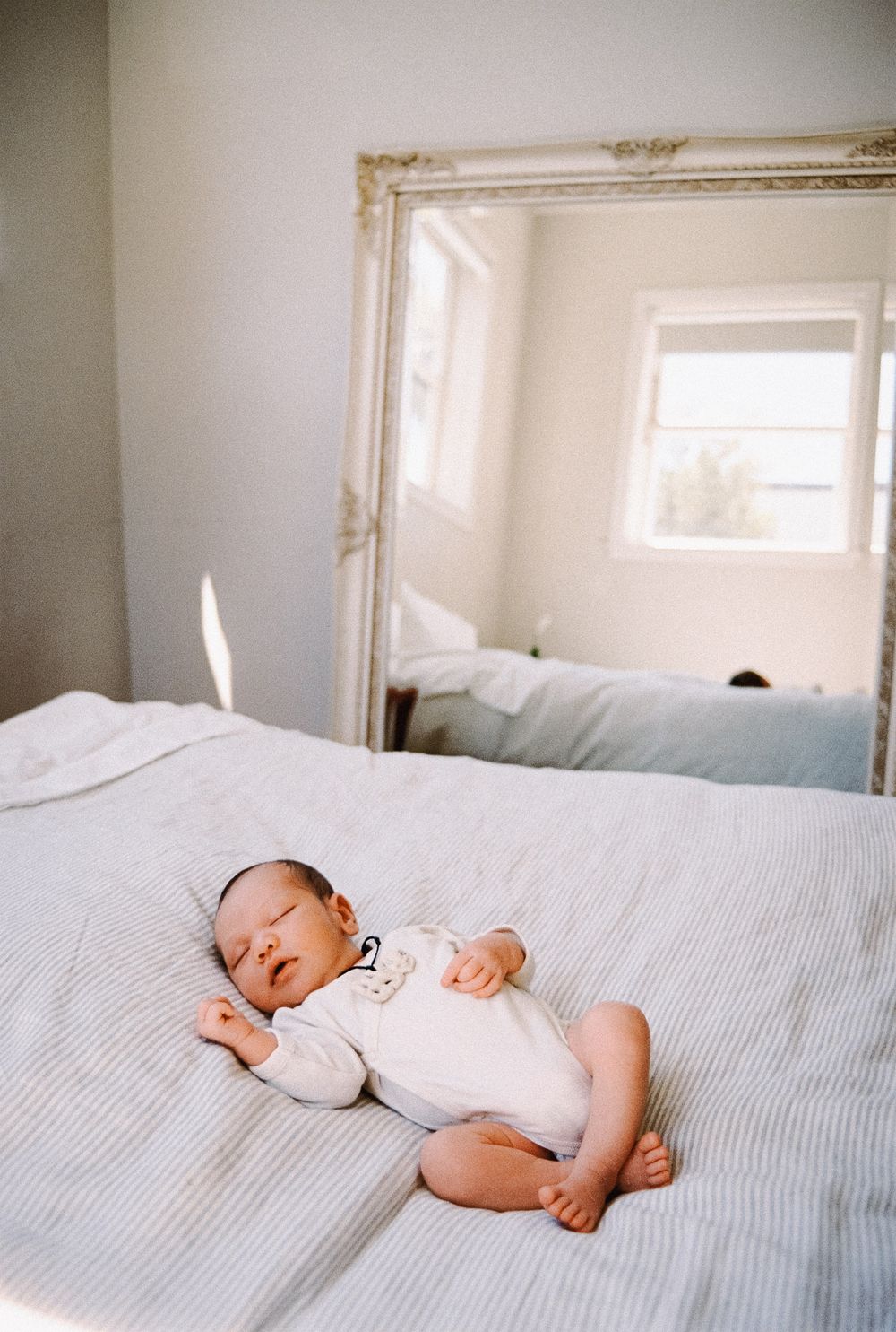 Peaceful newborn resting on a striped bed with vintage mirror and window in background.