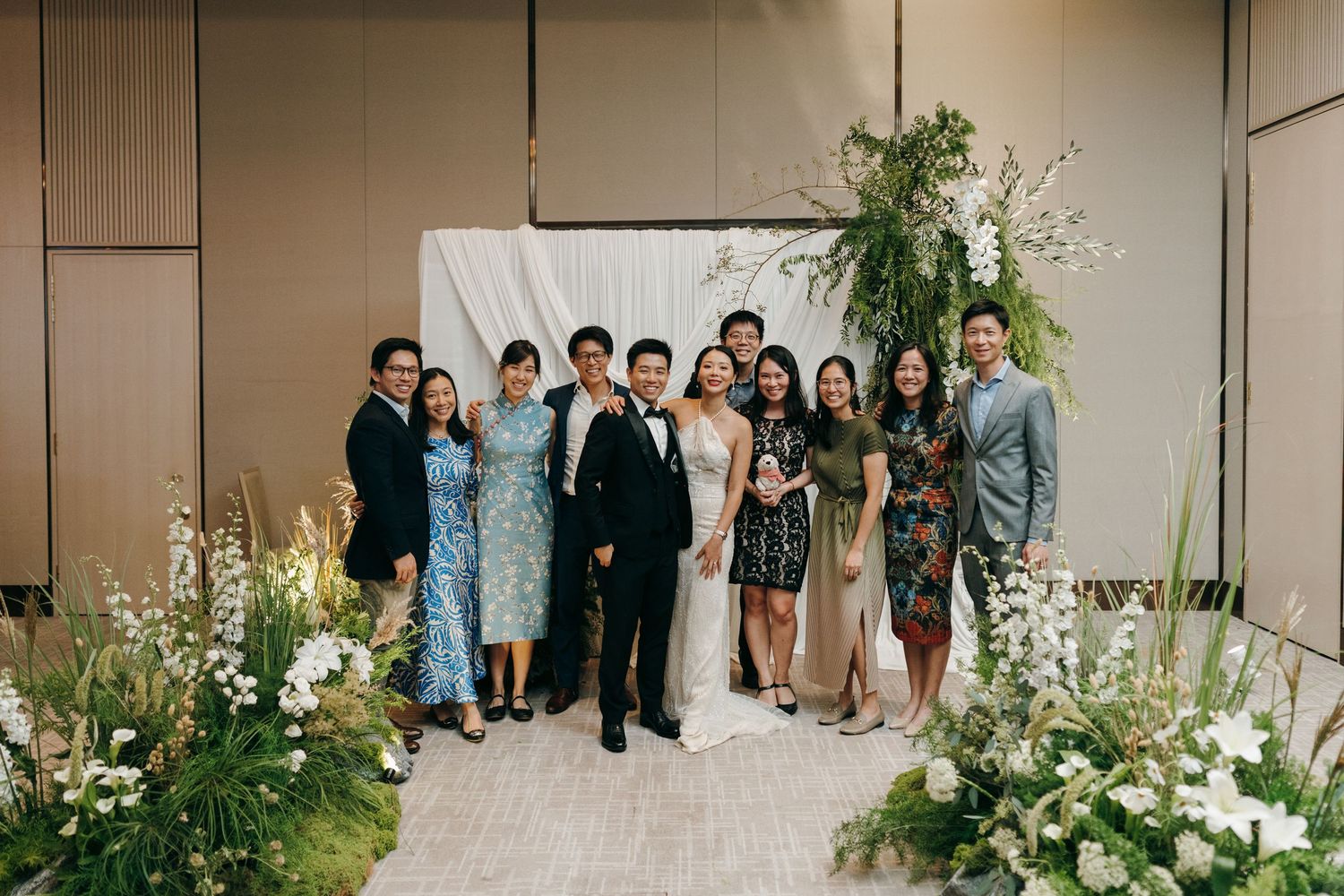 Group photo of wedding party members posing against a white backdrop with floral decorations.