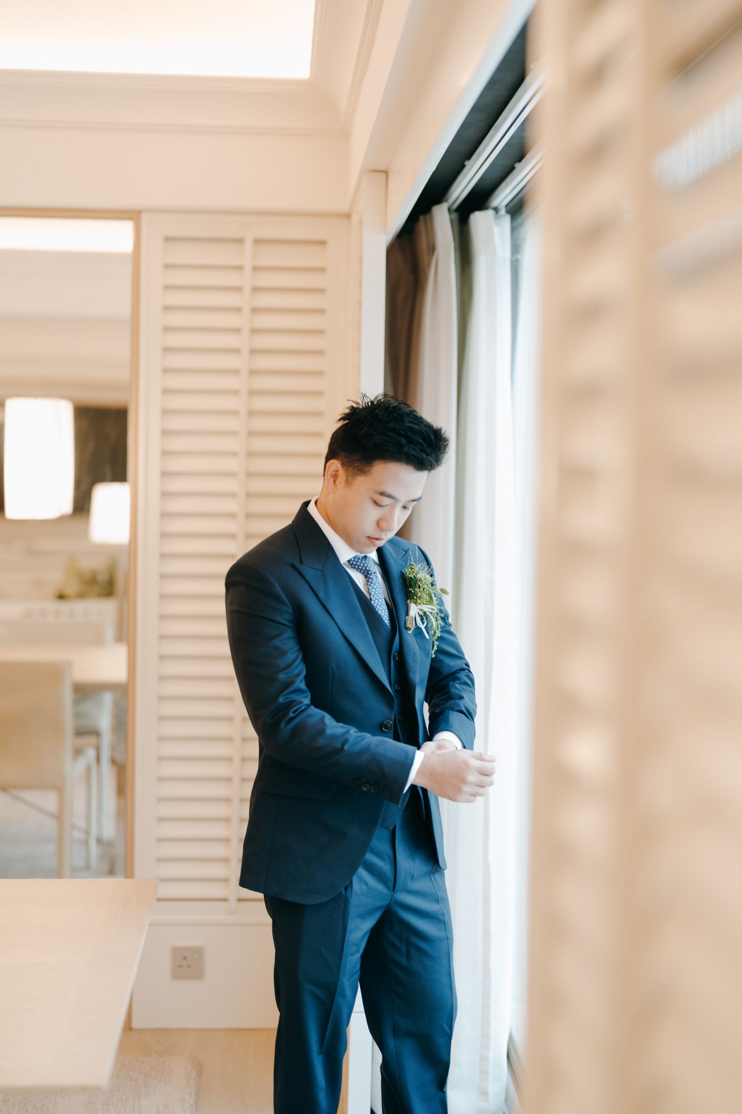 A person in a navy suit adjusts their tie while standing in a bright room with white shuttered windows.