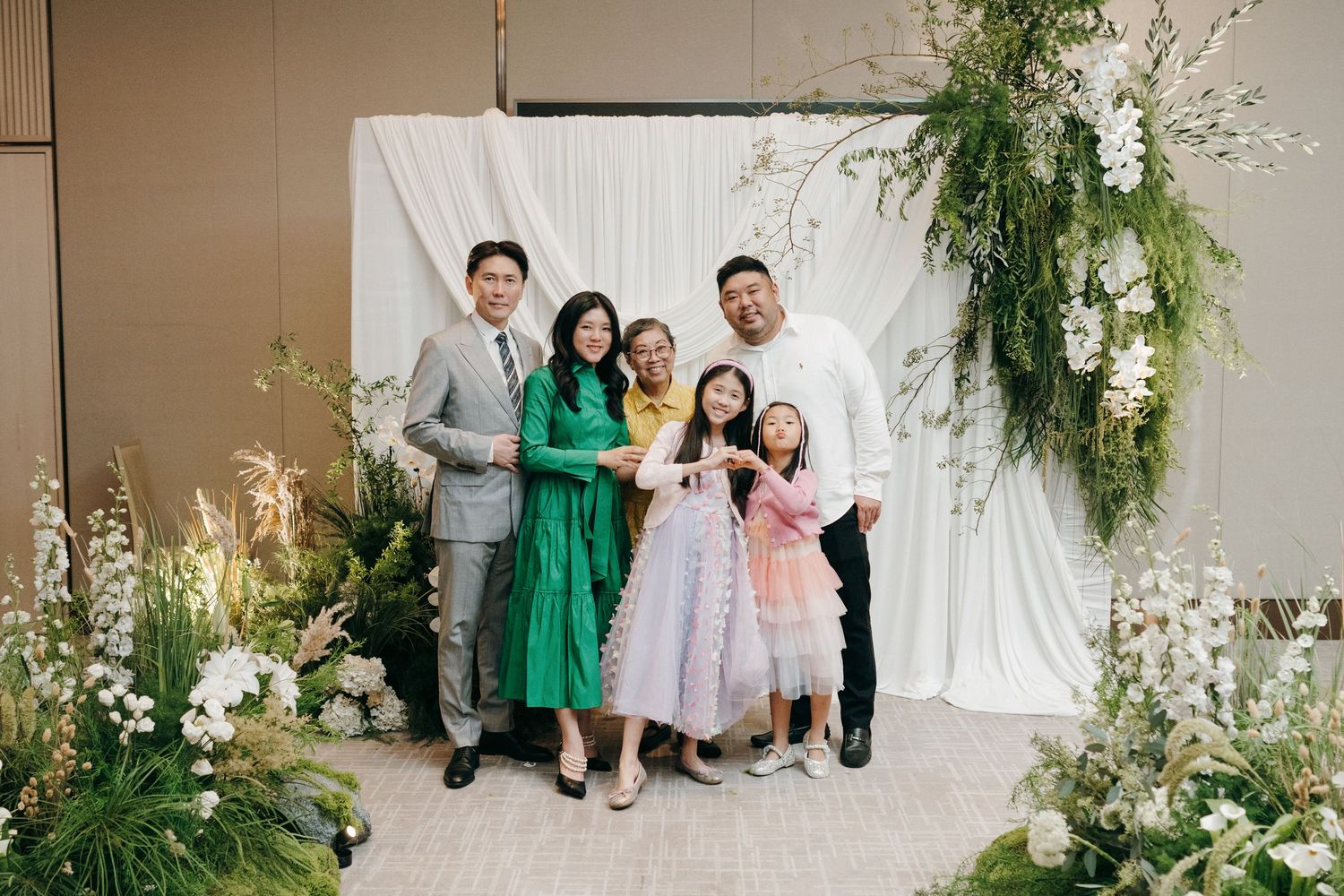 Group photo of wedding guests in formal attire posing against white backdrop with greenery decorations.