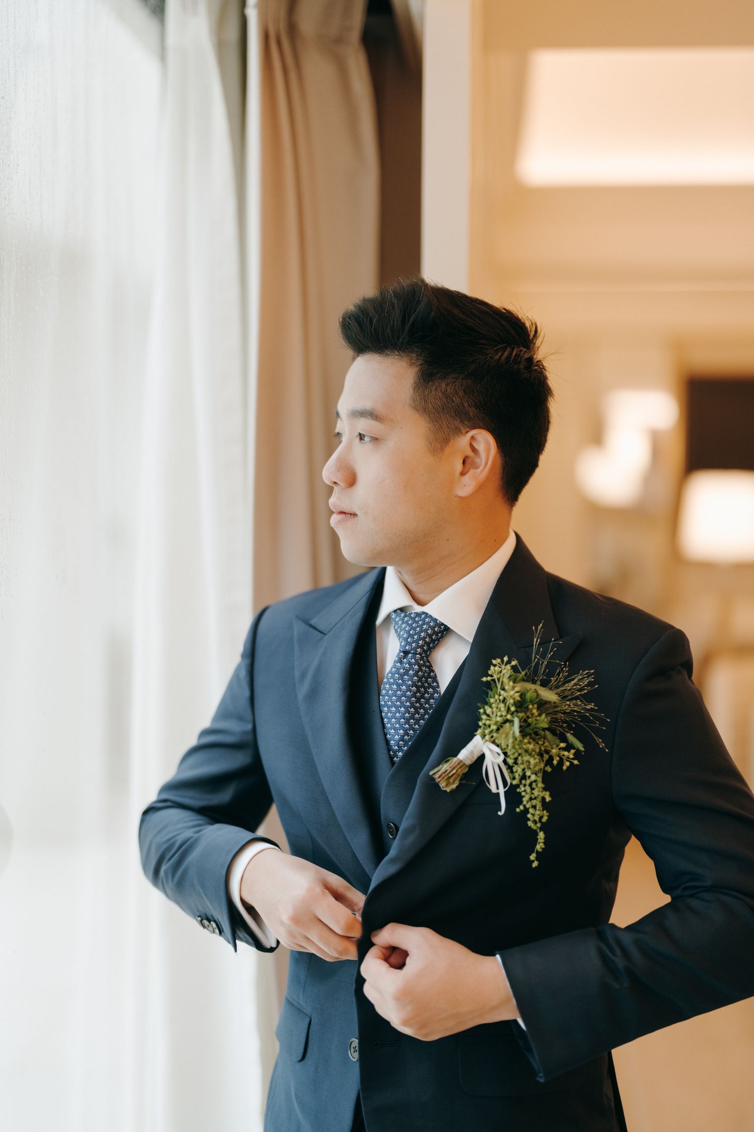 Groom in navy suit with boutonniere looking out window during wedding preparations.