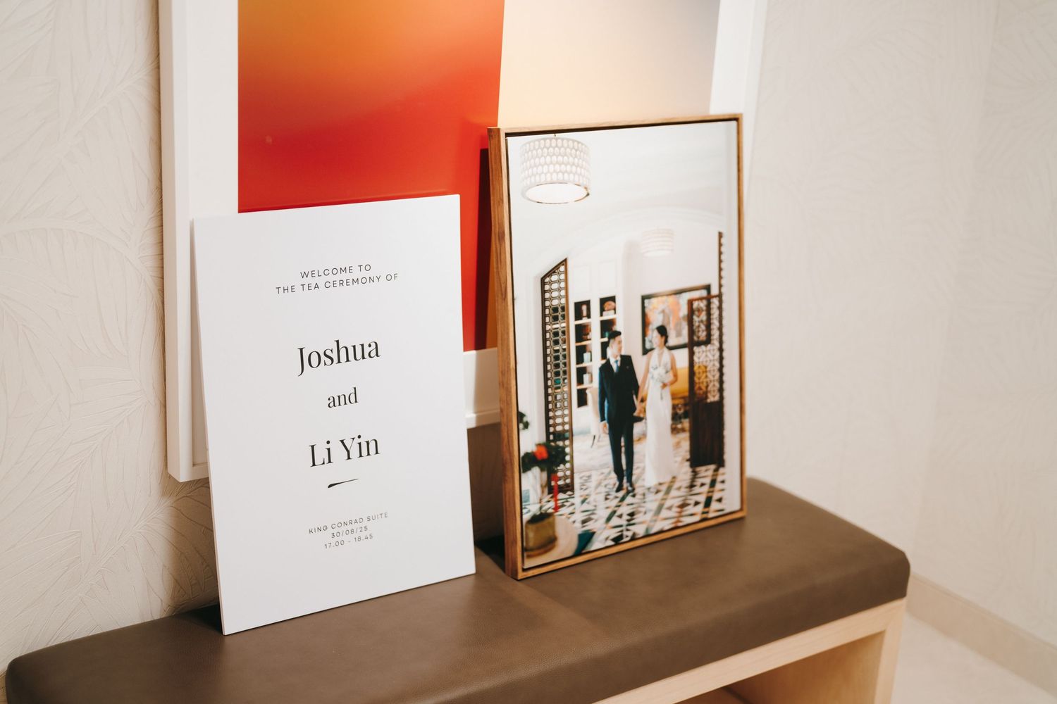 Wedding invitation and framed photo displayed on wooden surface with orange wall accent.