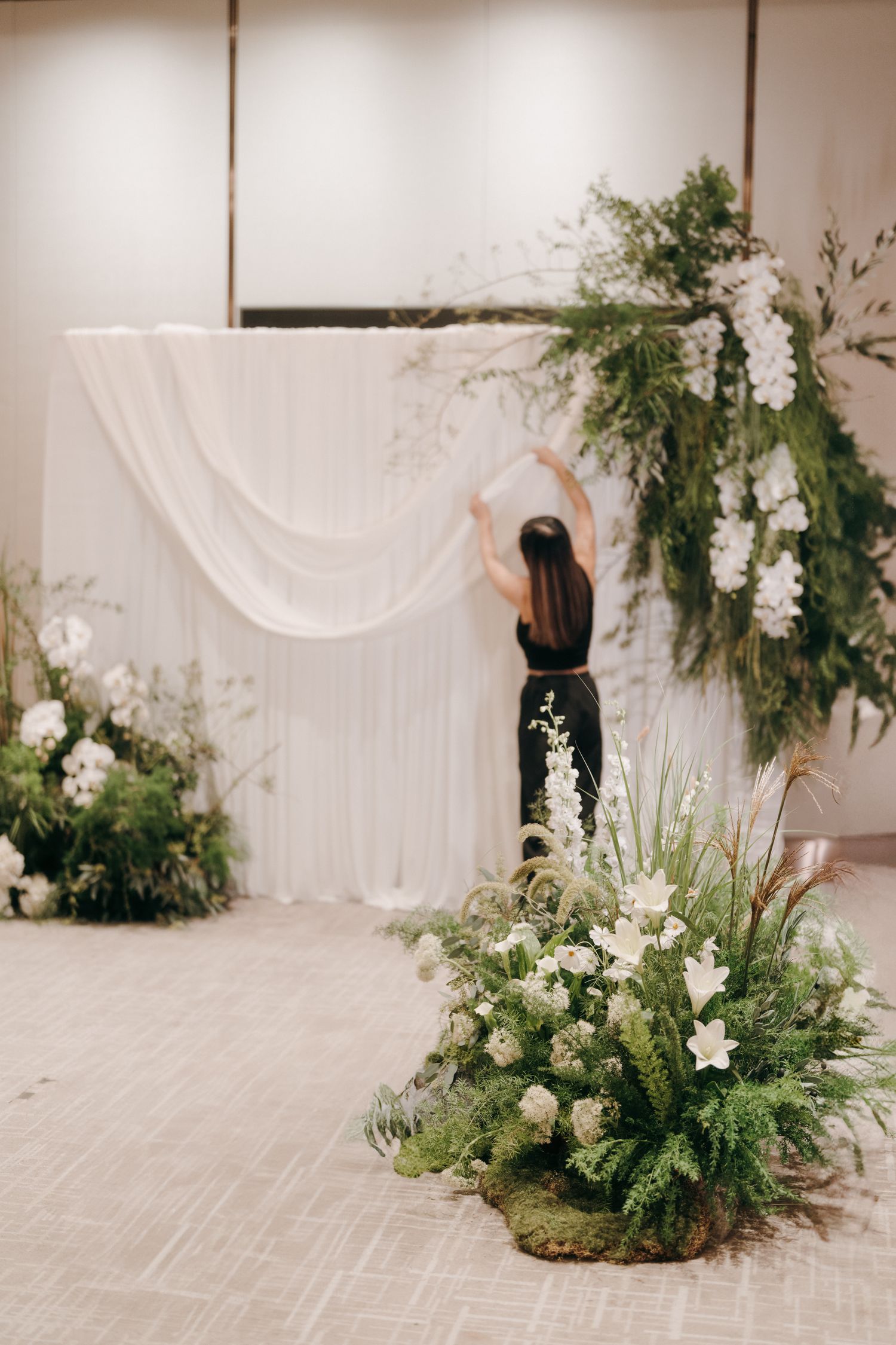 Modern wedding ceremony setup with white draping and lush greenery floor arrangements.