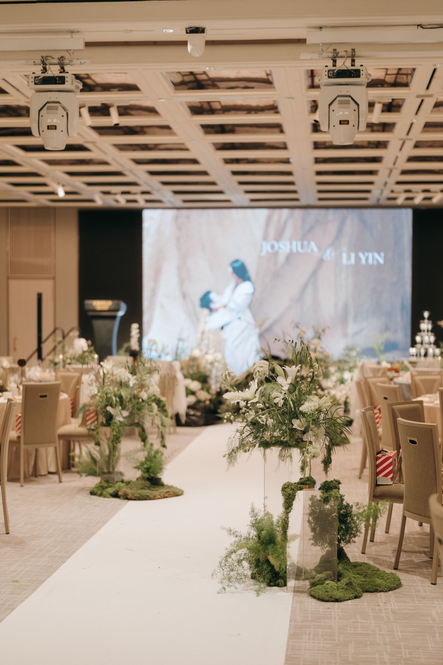 Wedding reception venue with minimalist decor, featuring floral centerpieces and projection screens.