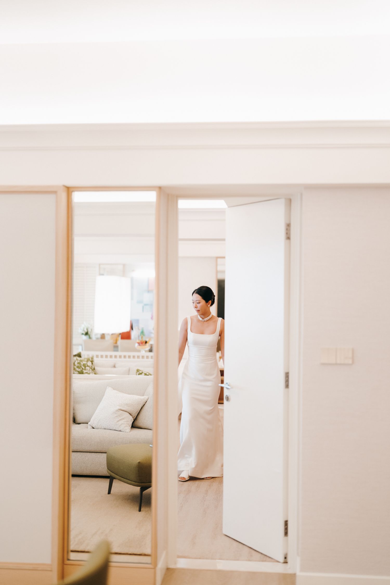 A reflection in a mirror shows someone in a white dress standing in a doorway of a modern room.