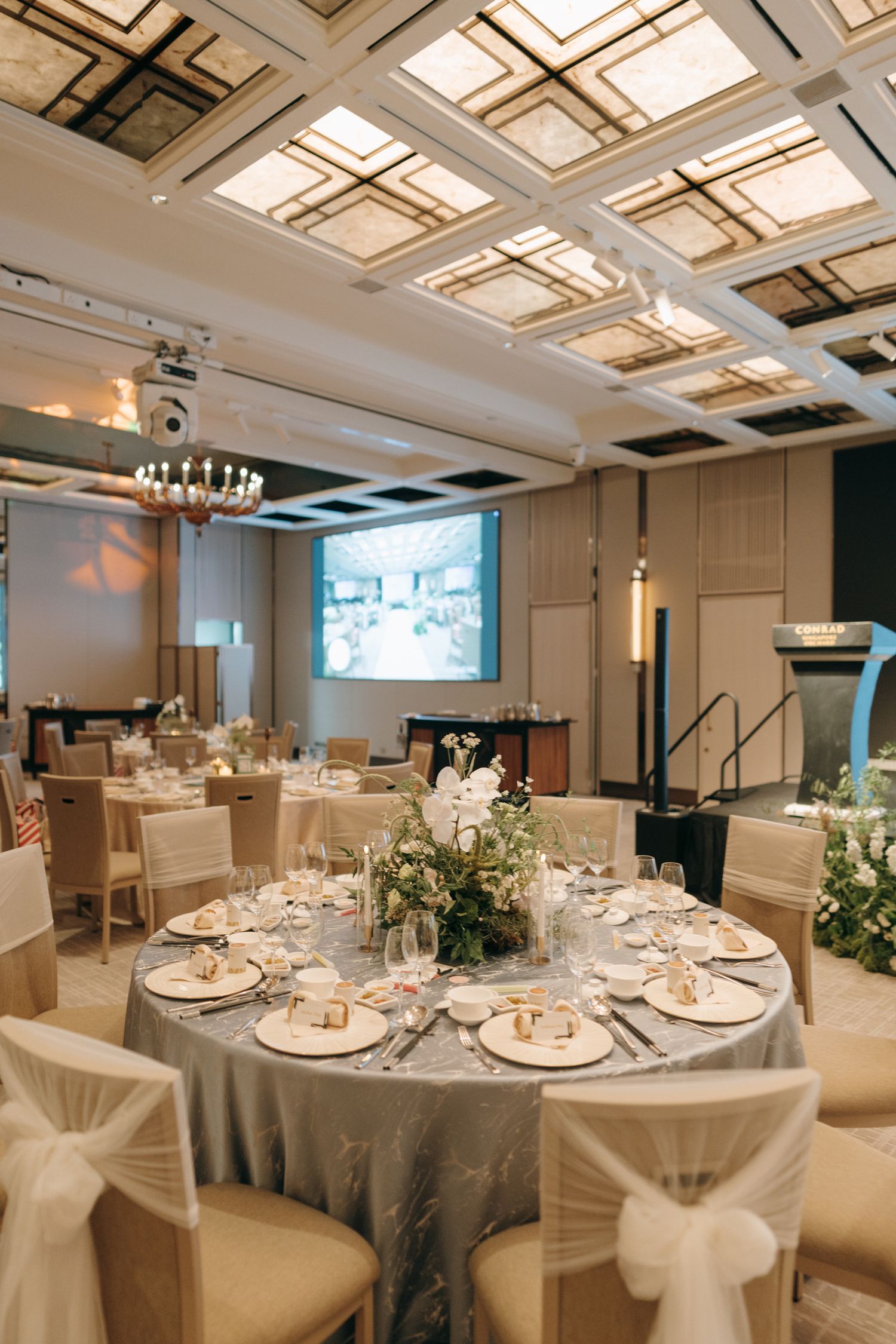 Modern ballroom setup with round tables, white chairs, and geometric ceiling details for an elegant reception.
