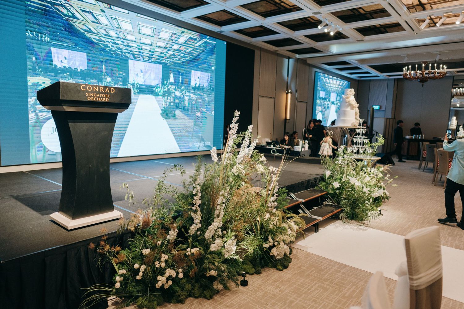 Wedding venue stage with podium and floral arrangements against a large projection screen.