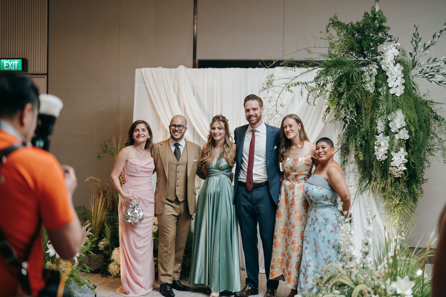 Wedding party posing in front of a white backdrop decorated with cascading greenery.
