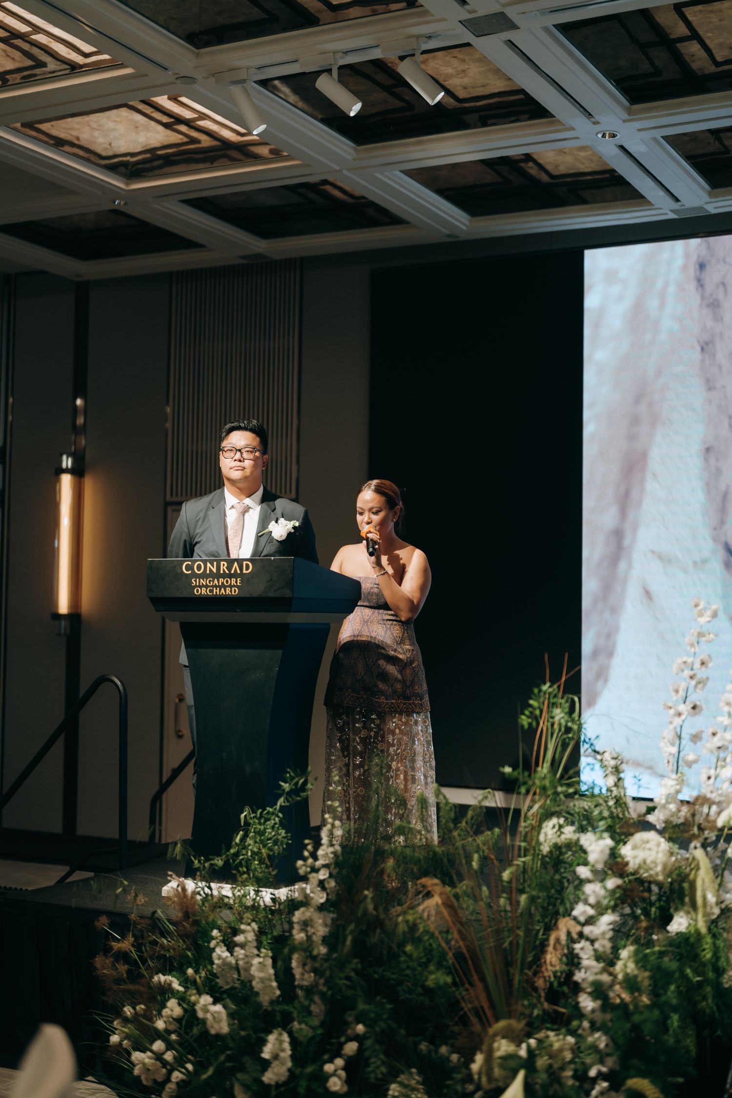 Speakers at wedding podium surrounded by white floral arrangements and projection screen backdrop.