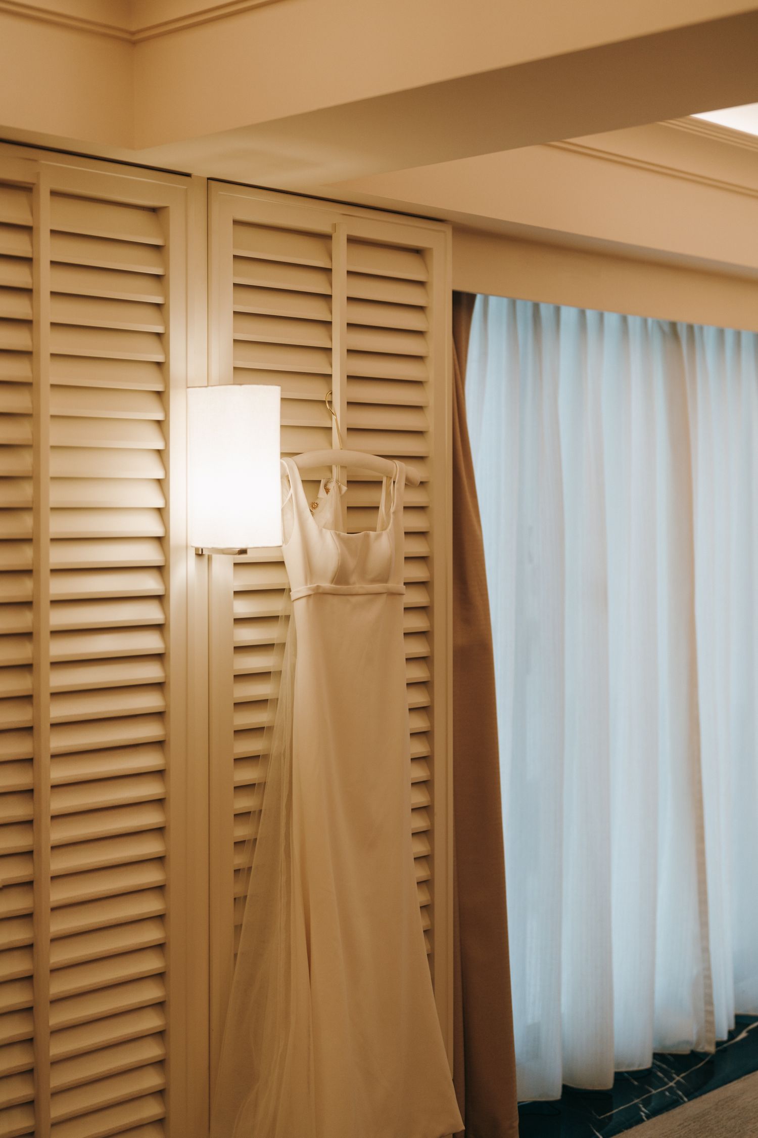 White wedding dress hanging on louvered doors next to a modern wall sconce.