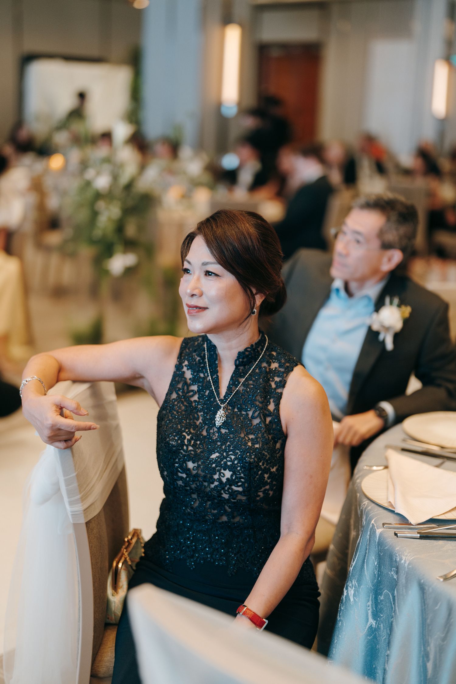 Guest in elegant black lace dress seated at formal reception table during wedding celebration.