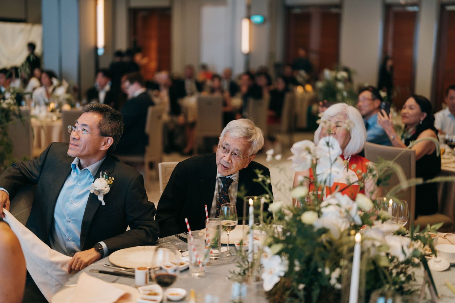 Wedding reception guests seated at round tables with elegant floral centerpieces in an indoor banquet hall.