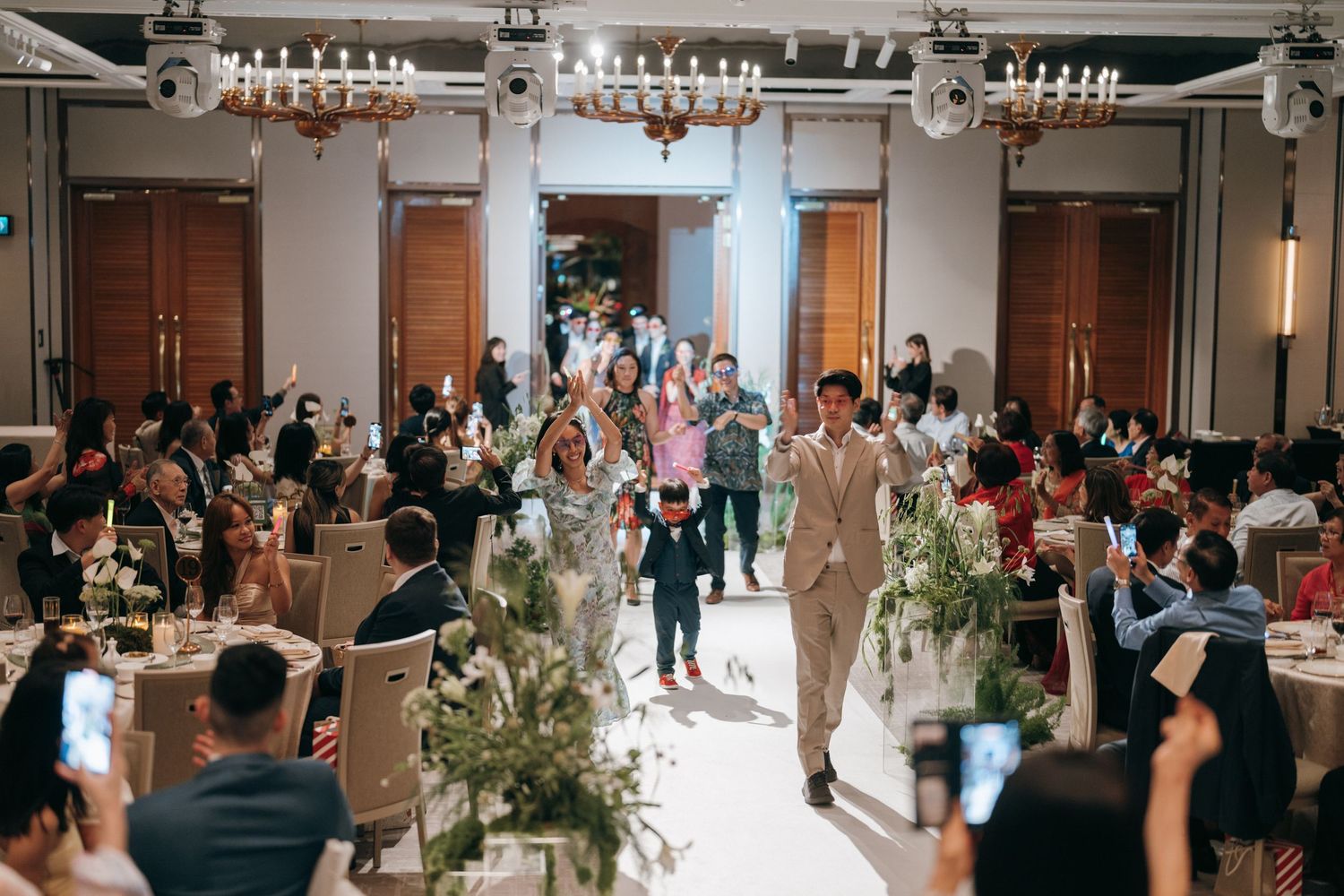 Wedding reception guests gather in an elegant banquet hall with floral decor as a couple walks down the center aisle.