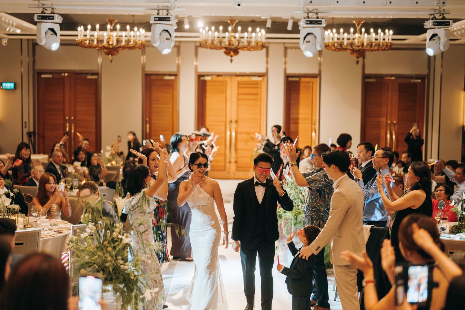 Guests dancing and celebrating at an elegant wedding reception in a grand ballroom with ornate decor and floral arrangements.