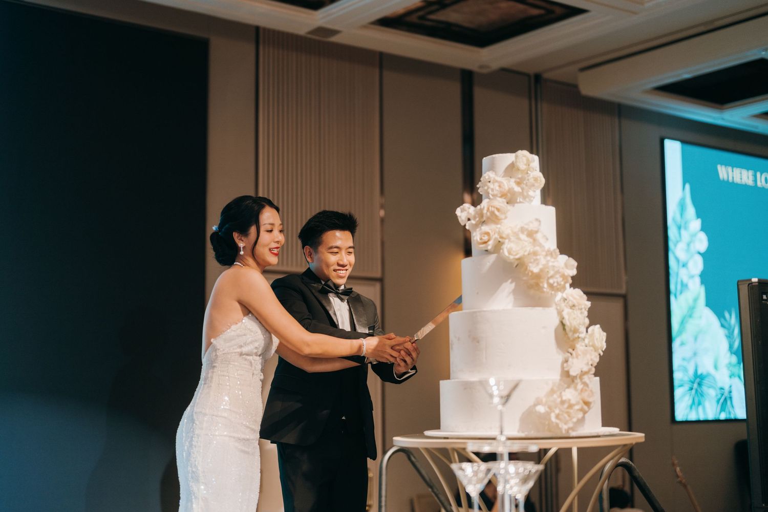 A couple cuts into their elegant white wedding cake adorned with cascading floral details at their reception.