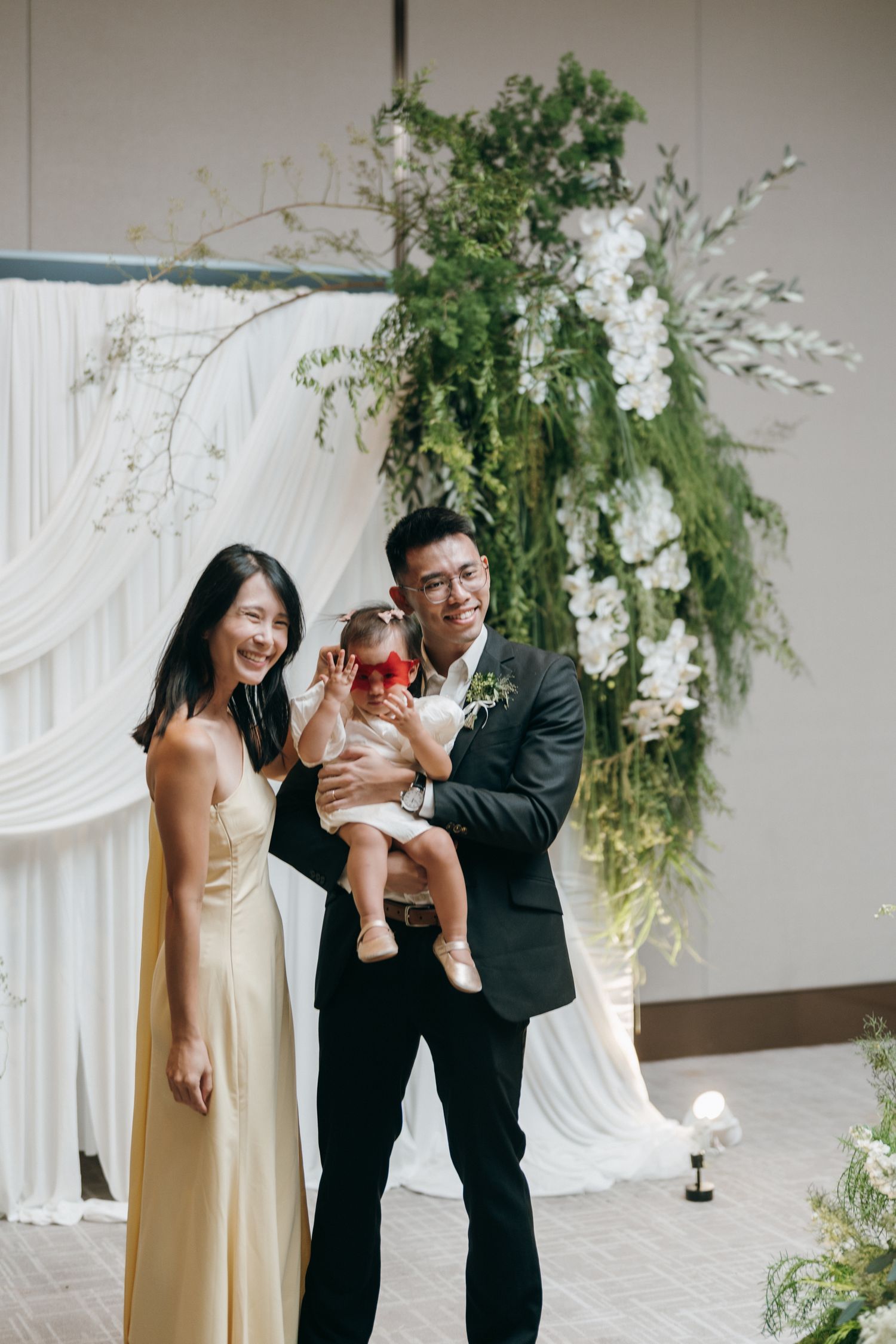A family shares joyful moments together in a white-draped event space decorated with elegant greenery arrangements.