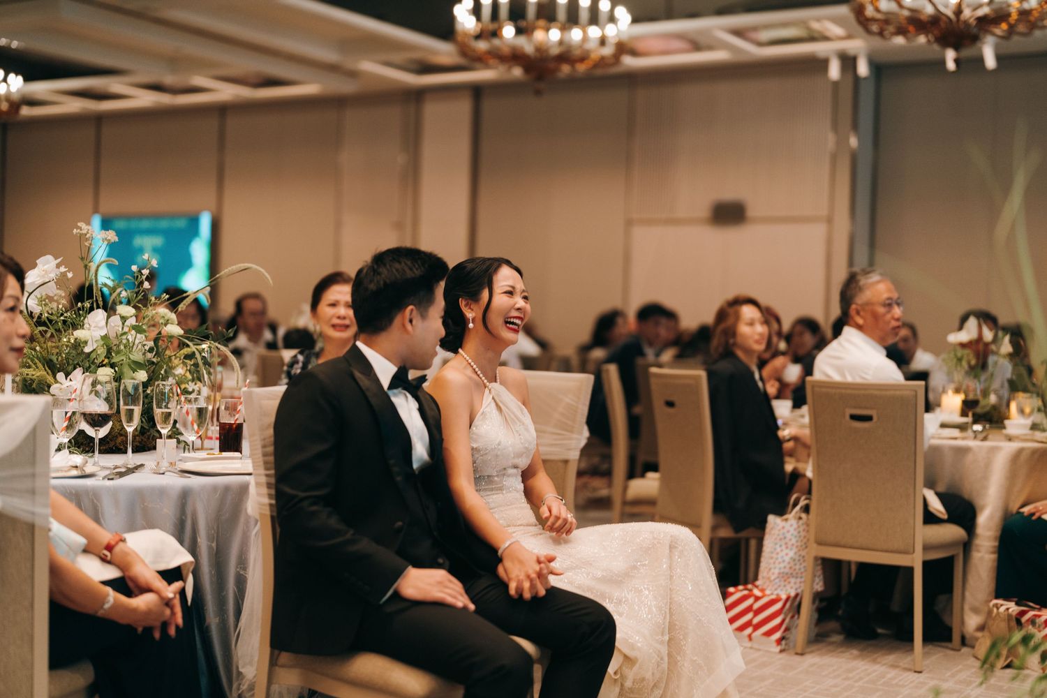 Wedding guests seated at round tables in a ballroom with chandeliers and elegant decor.