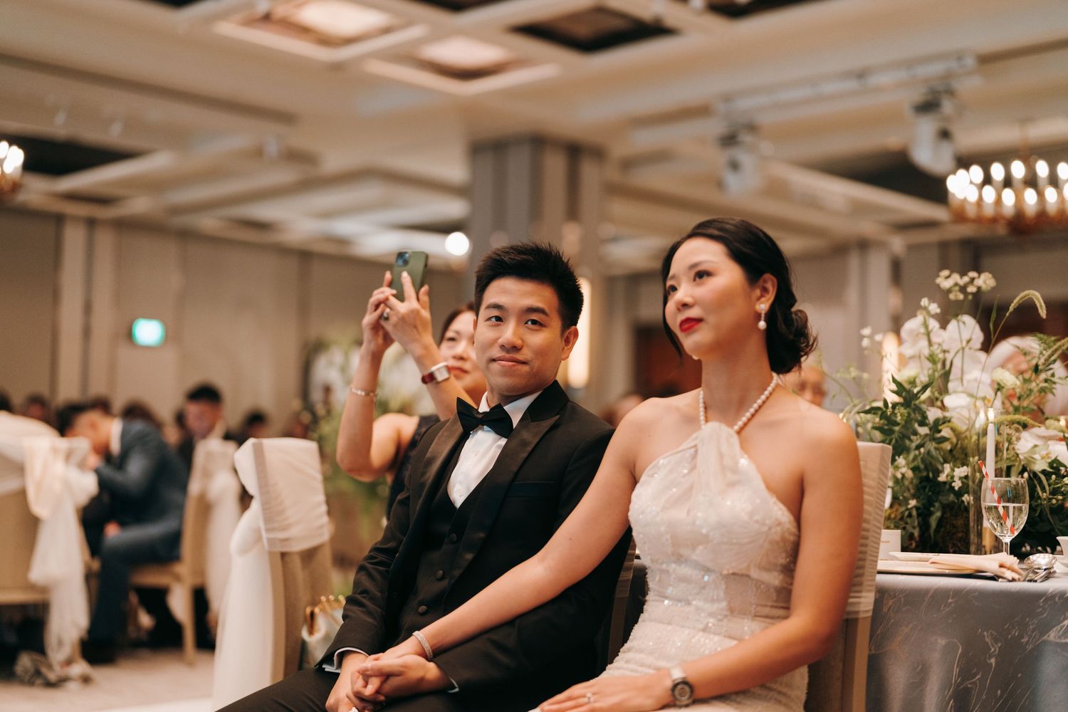 A couple in formal attire celebrates at an elegant wedding reception with champagne glasses raised in a ballroom.