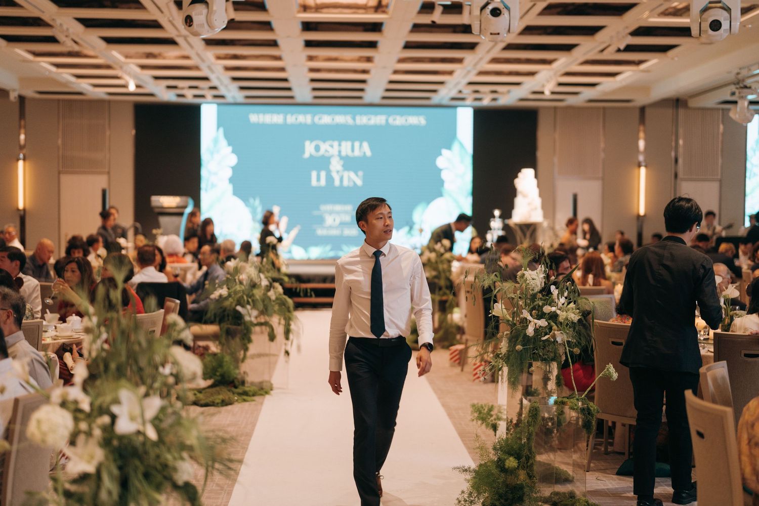 Wedding reception venue with floral decorations and a white backdrop screen displaying event details.