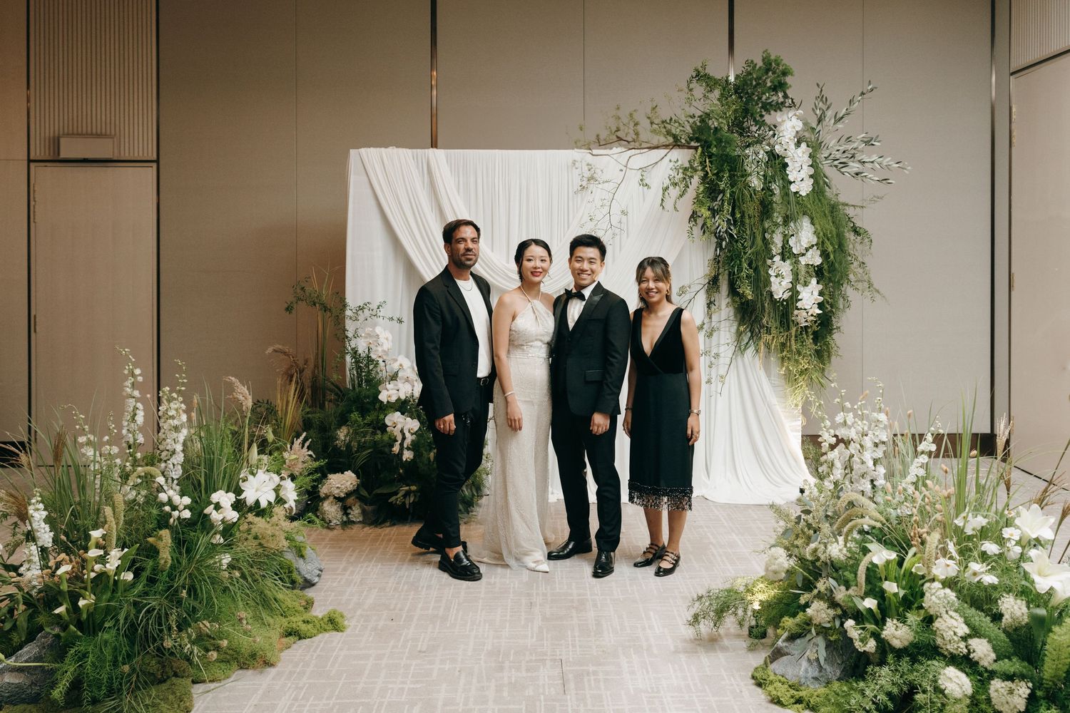 Wedding party poses in front of elegant white backdrop with cascading greenery and floral arrangements.