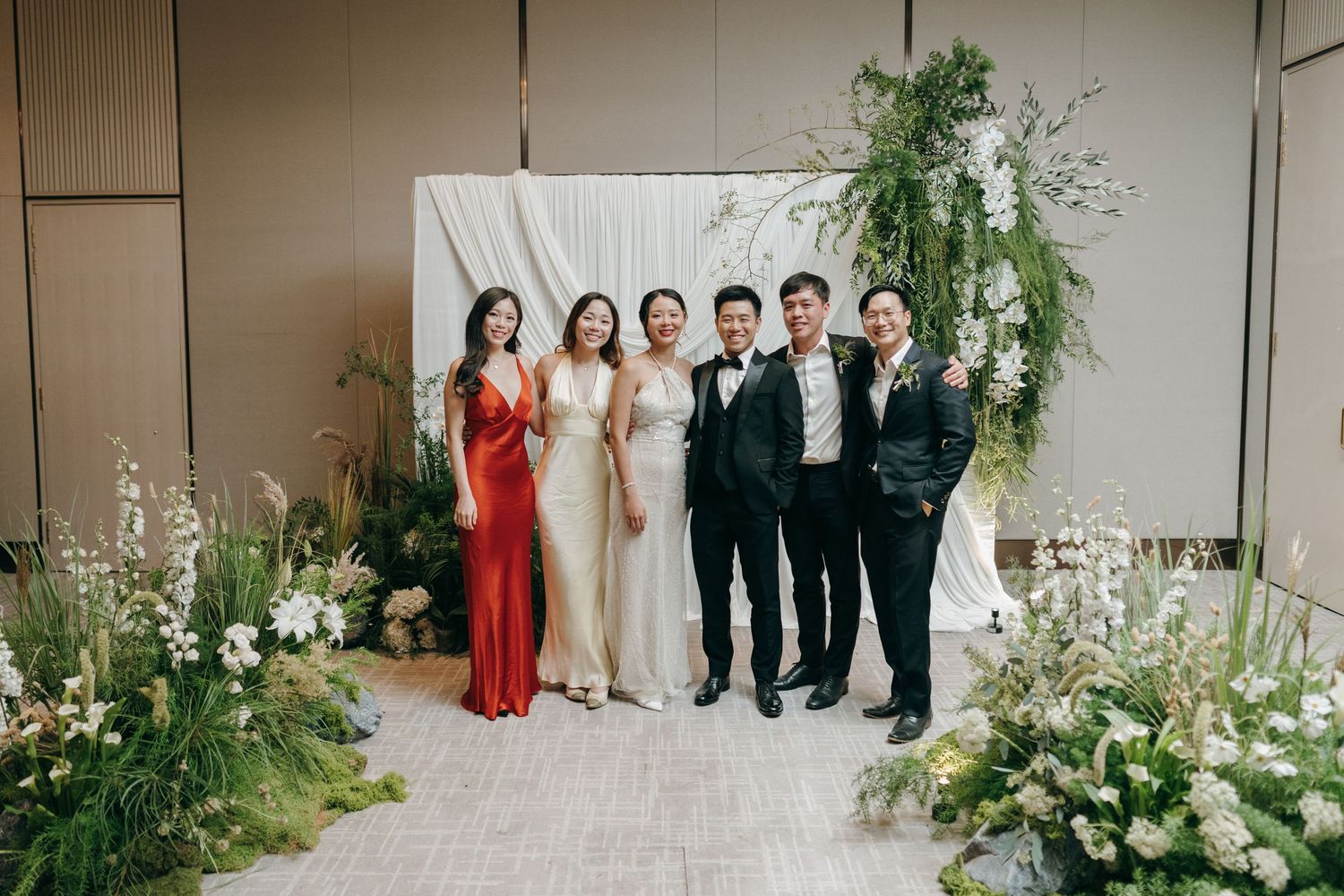 Wedding party poses for formal photos with elegant floral arrangements and white backdrop.