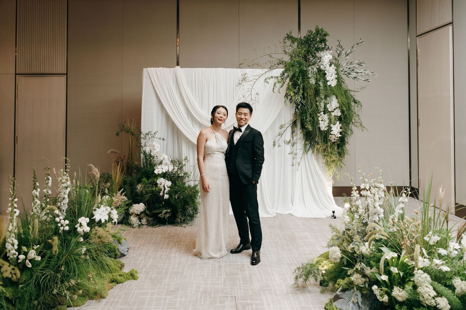 Couple poses in front of white draped backdrop with elegant floral arrangements at wedding venue.