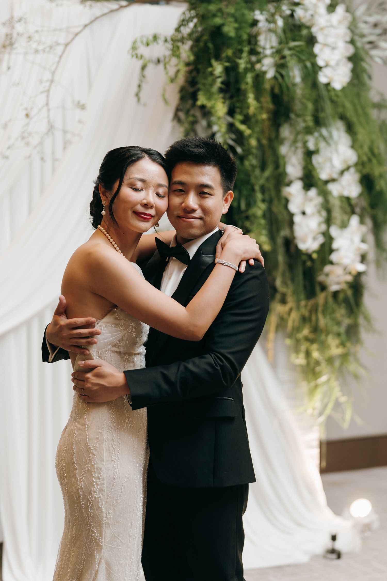 Elegant wedding couple embraces in front of white drapery and hanging floral arrangements.