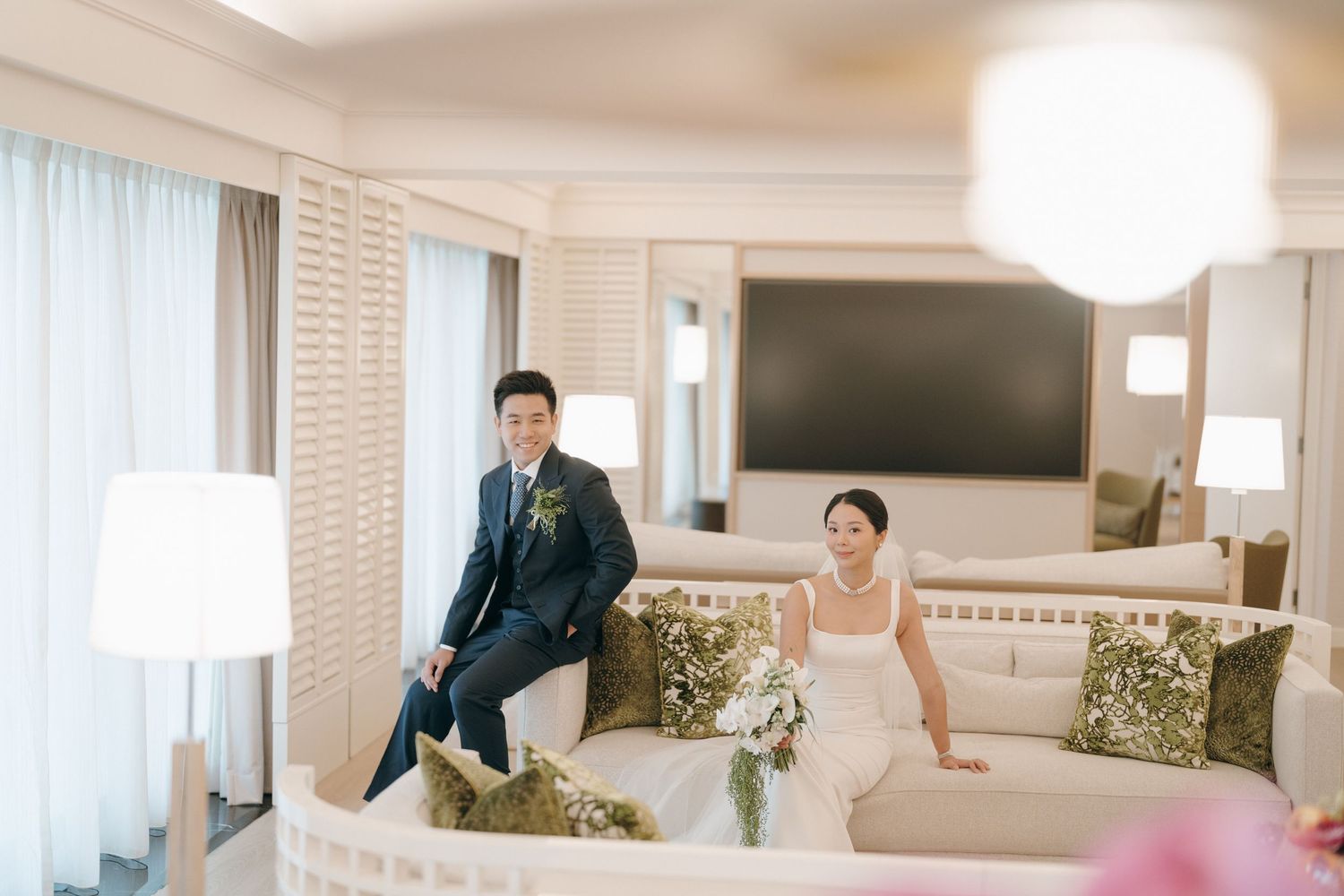 A wedding couple sitting casually on opposite ends of a luxurious hotel room sofa.