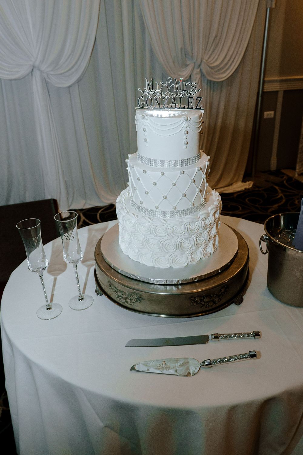 Elegant three-tier white wedding cake with pearl details displayed on silver cake stand with champagne flutes nearby.