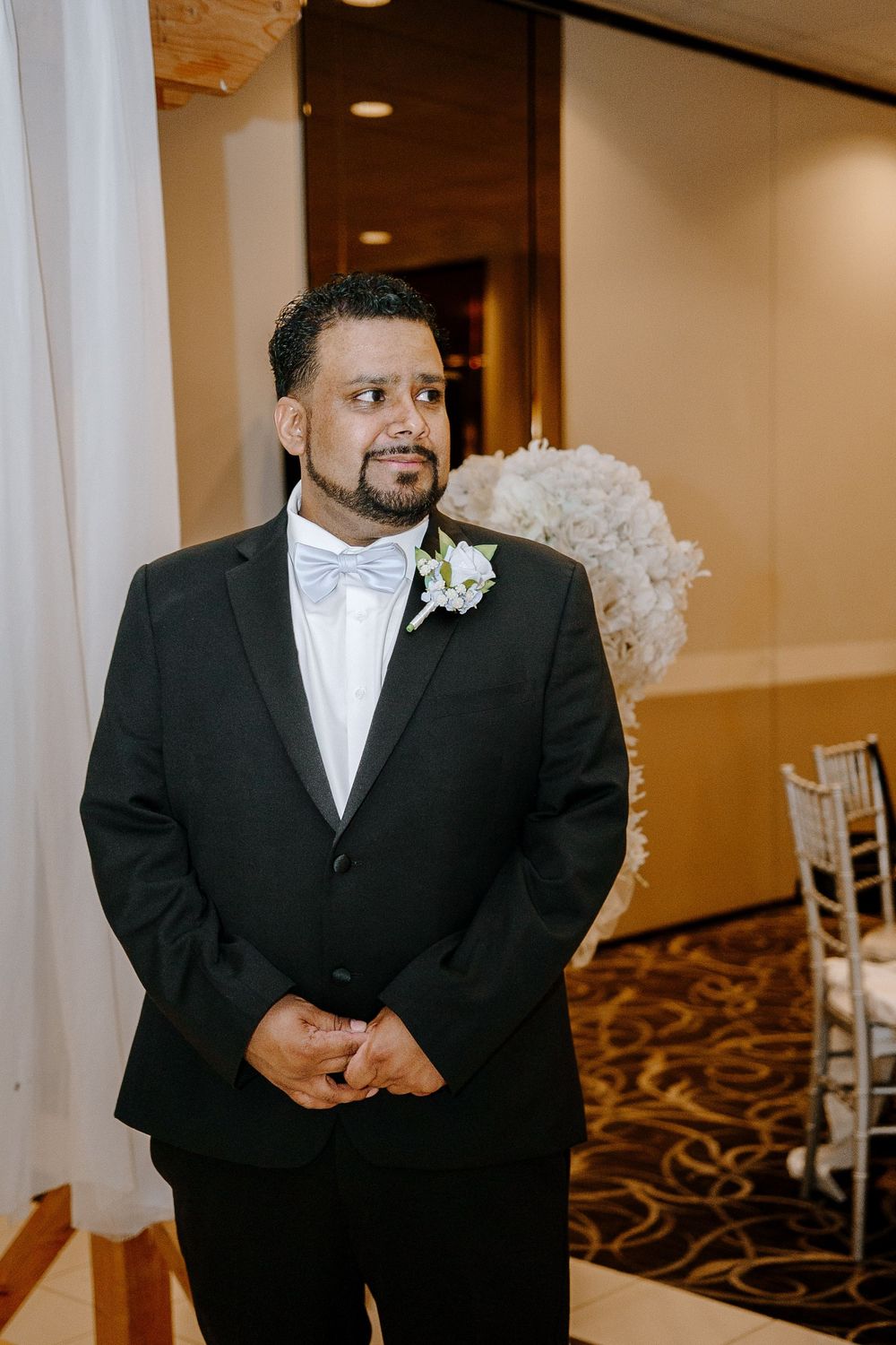 A person in a black tuxedo with white bowtie smiles during a formal event in an elegant venue.