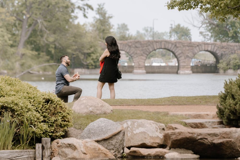 A romantic proposal scene on a rocky shore with a stone bridge in the background near a serene lake.