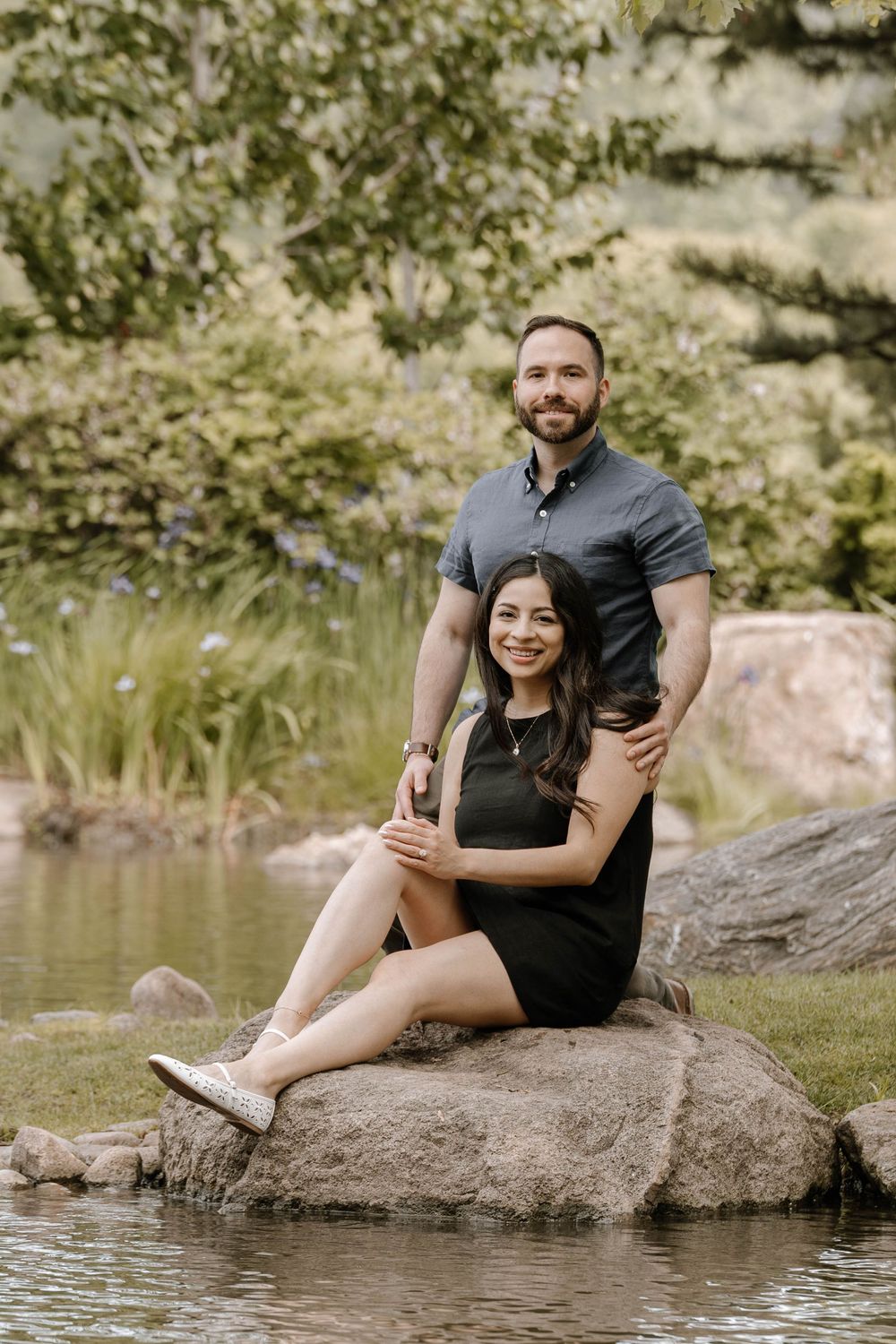 A couple sits intimately together on a large rock by a serene pond in a natural outdoor setting.