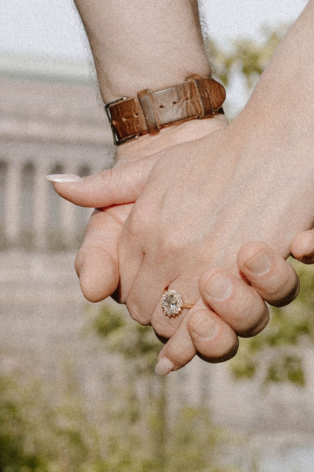 Close-up of two hands holding each other with a diamond engagement ring visible in a romantic outdoor setting.