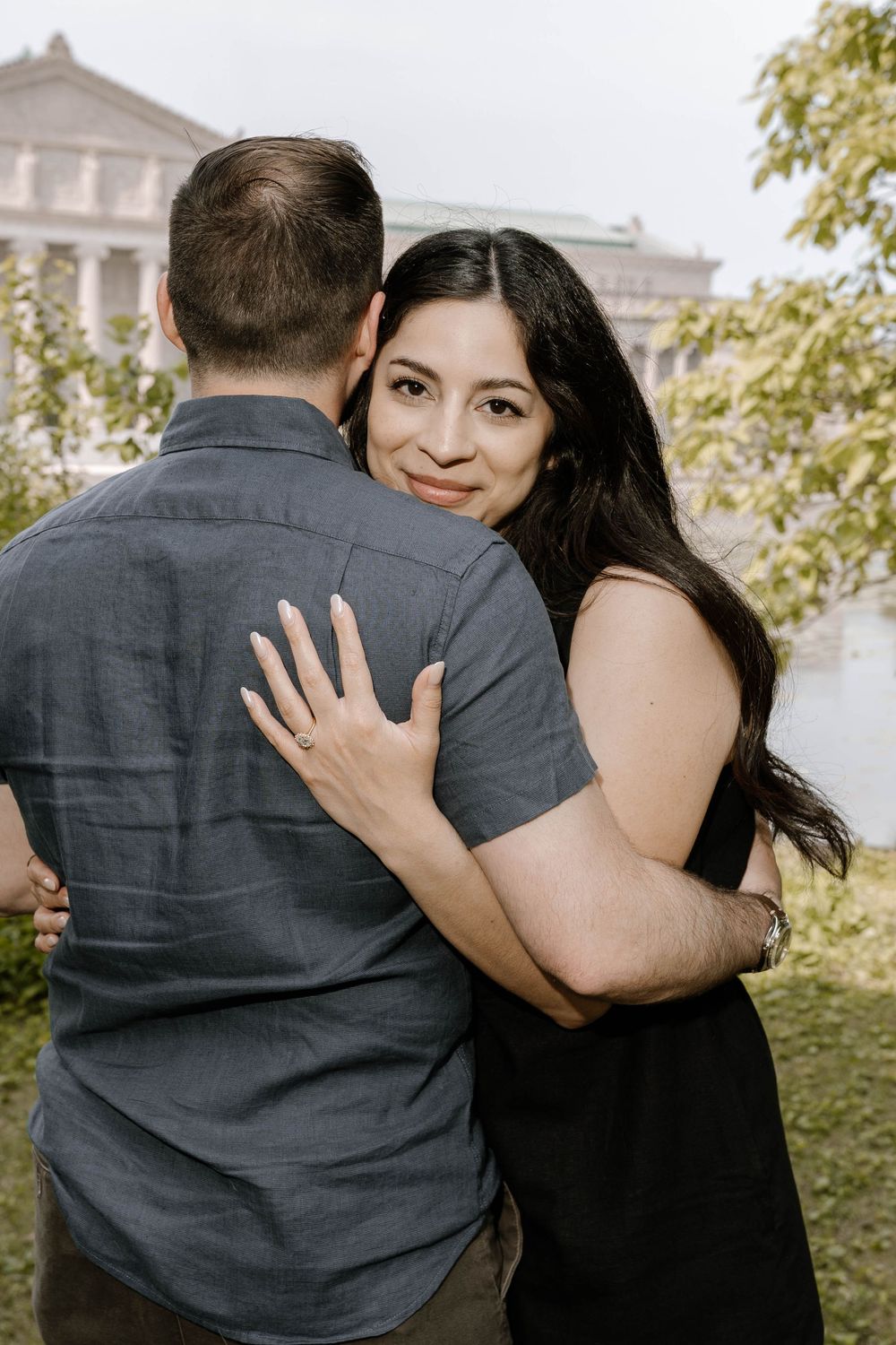 A couple in a black dress and gray suit embrace outdoors with architectural elements visible in the background.