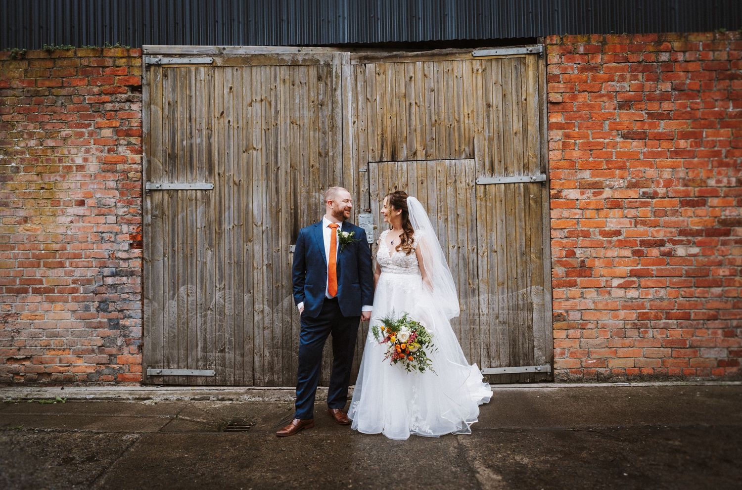 A couple in wedding attire poses against rustic brick walls and wooden doors in a romantic outdoor setting.