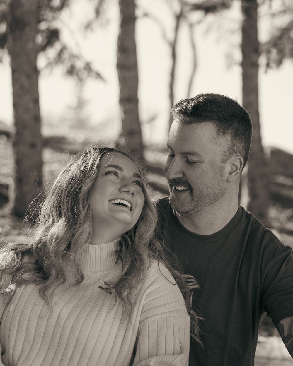 Black and white photo of a laughing couple sharing a joyful moment outdoors among trees.