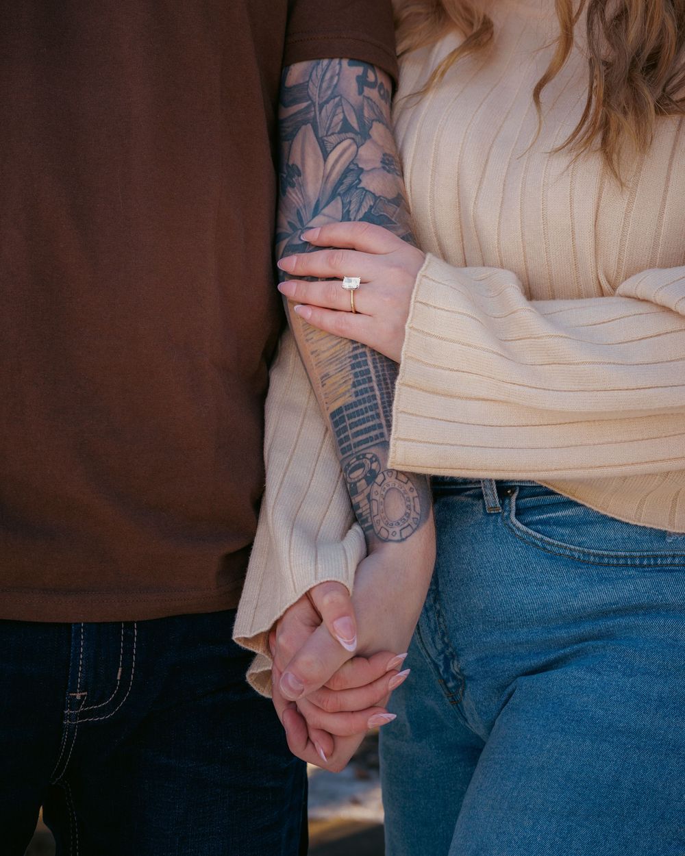 Close-up of an engaged couple holding hands, showcasing a diamond ring and a tattooed sleeve.