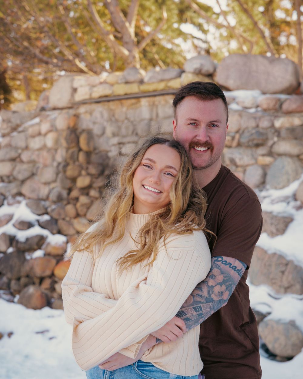 Happy engaged couple posing together in a snowy winter setting with a stone wall backdrop.