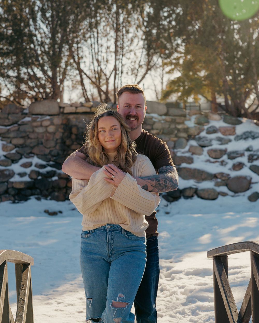 Couple embracing warmly in a snowy outdoor setting with golden sunlight and a stone wall behind.