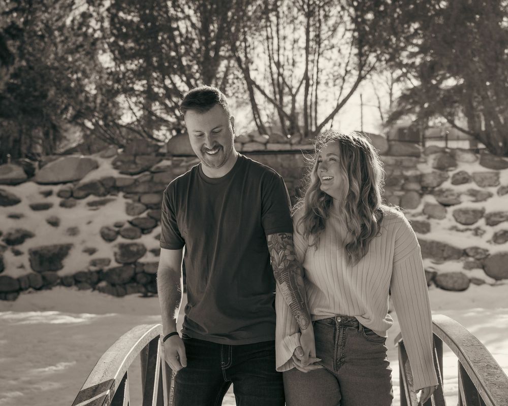 Black and white photo of a laughing couple walking together on a bridge in a snowy park.